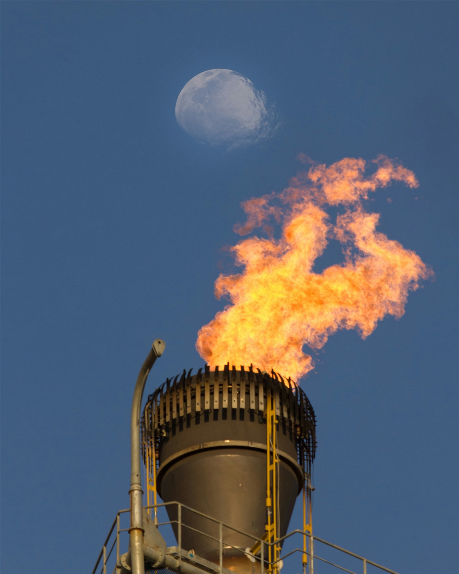 Photo by Matin Tavazoei Gas flare burning against a blue sky with the moon overhead